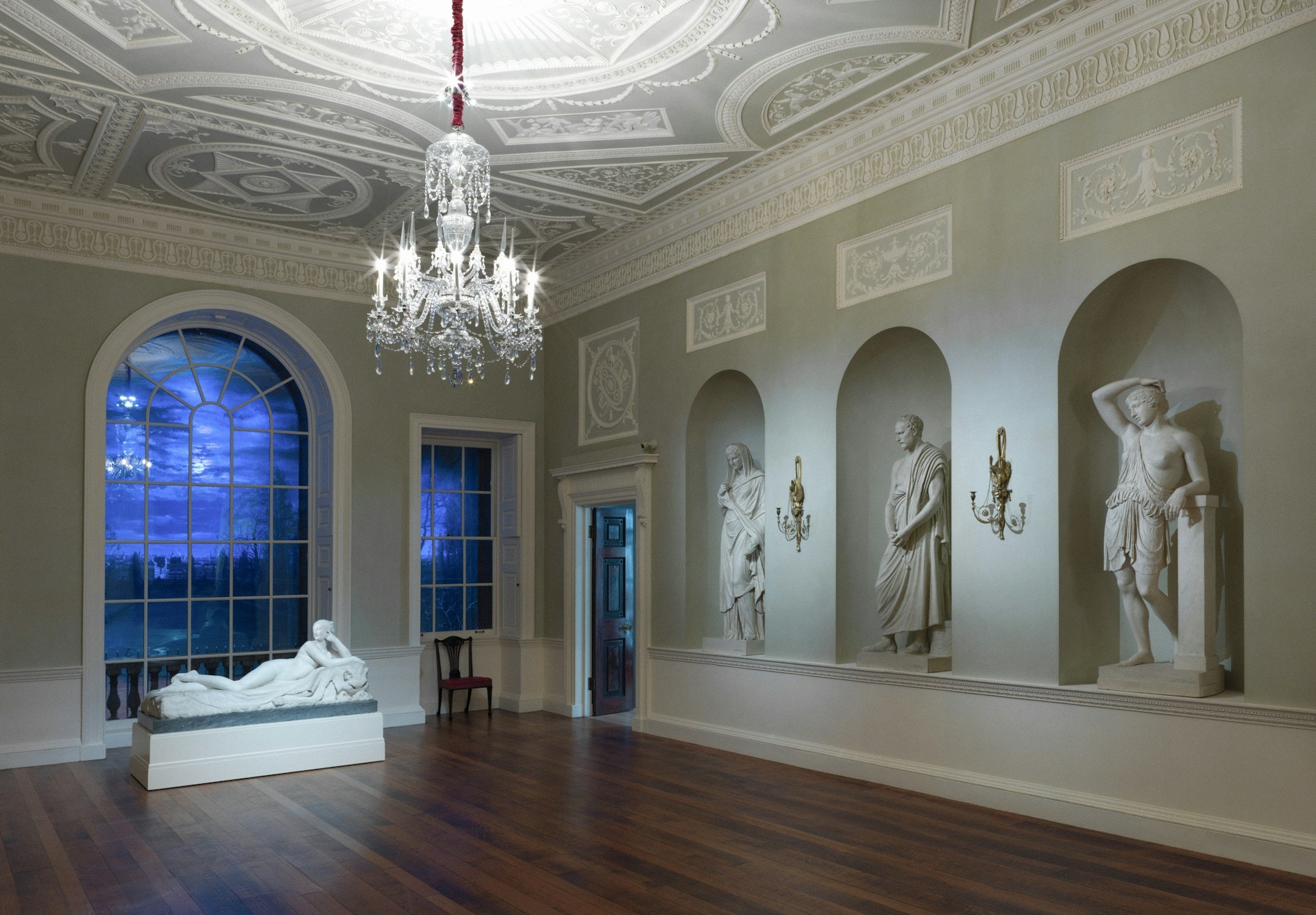 Et statuegalleri med hvide marmorstatuer i buede nicher og en lysekrone foran et måneoplyst vindue