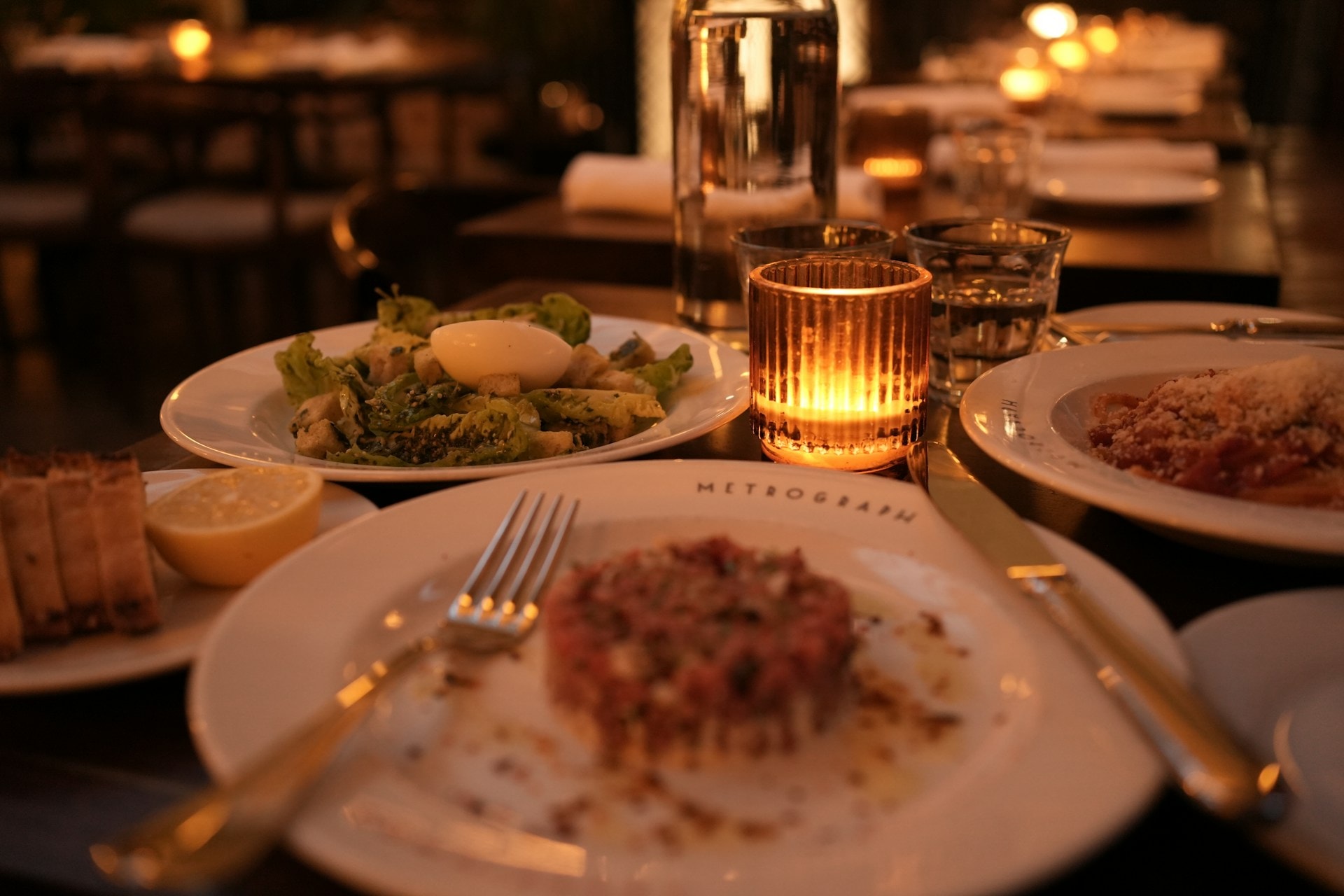 Et stearinlysbelyst restaurantbord med anretninger, glas og en vinflaske i baggrunden