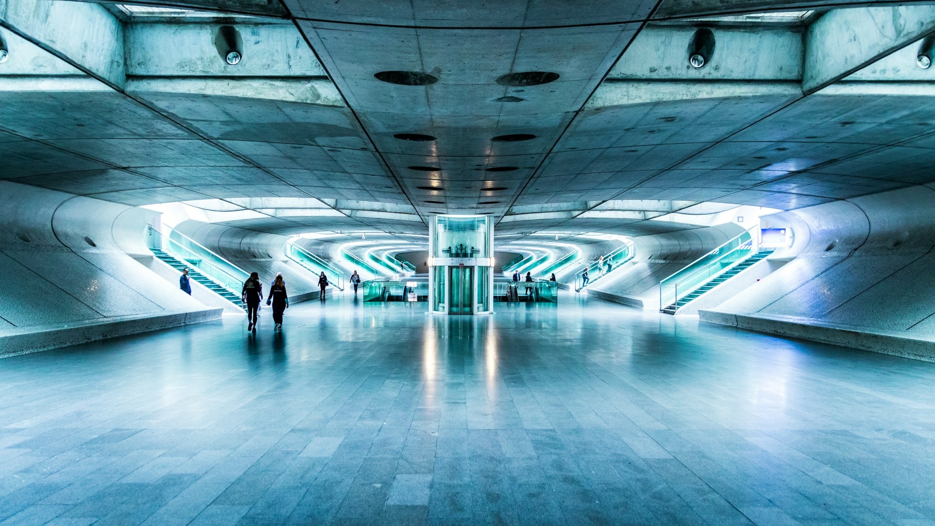 Mennesker går som skygger gennem en futuristisk, symmetrisk stationshal i koldt blåt lys