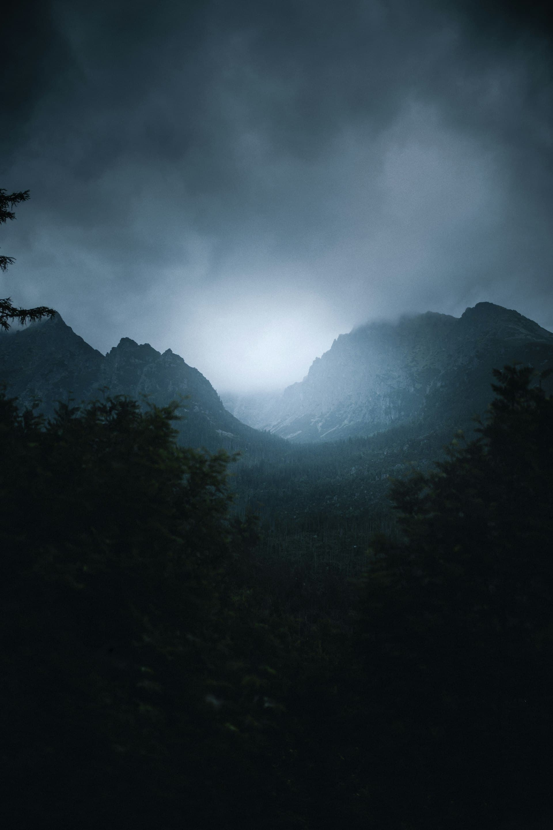 Misty mountains shrouded in dark clouds at dawn, dramatic layers of fog rolling through dark forested ridges